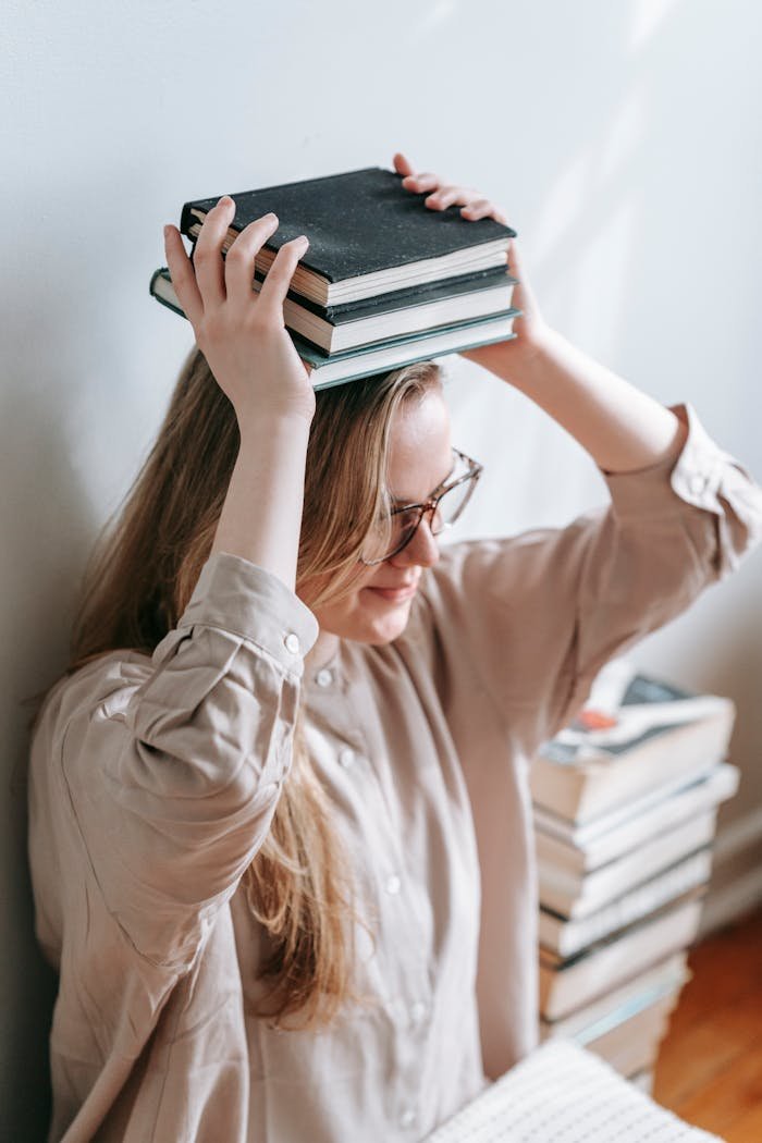 High angle of female in eyeglasses sitting near wall and putting books on head for improving balance