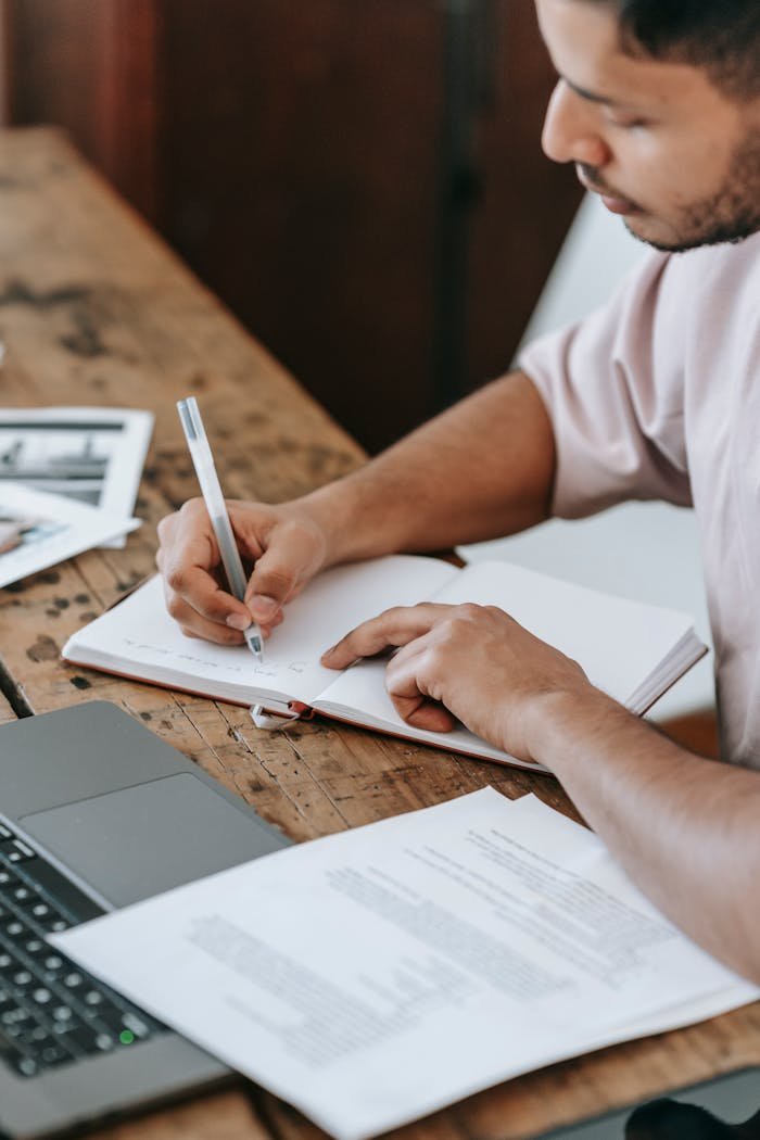 Crop concentrated male taking notes in notebook while sitting at desk with netbook and studying